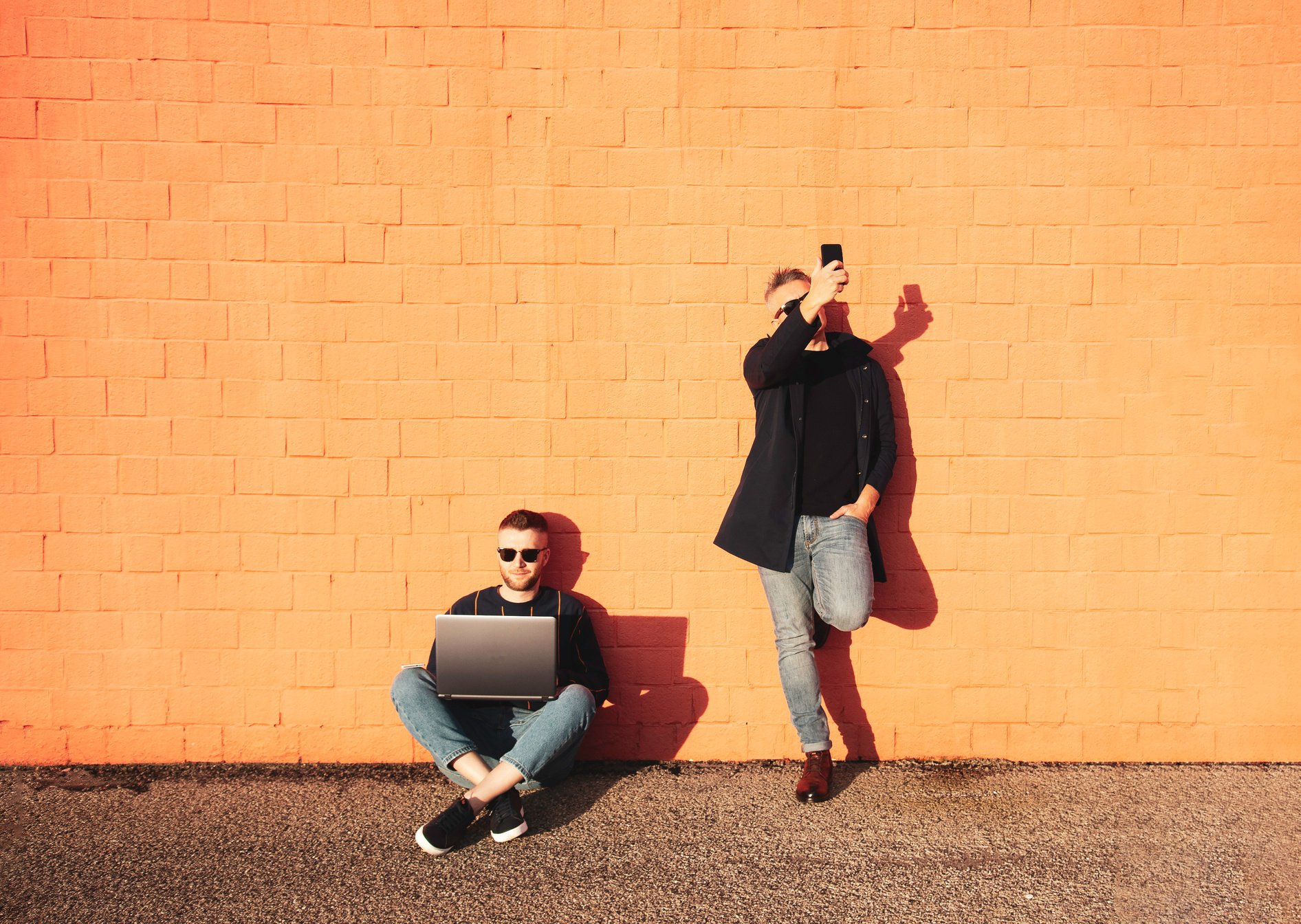 Two Friends or Colleagues with Digital Devices during Lunch Break - Young Caucasian Man Sitting Cross-Legged on Ground and Working on Laptop - the Other Man Standing and Taking Selfie with Smartphone